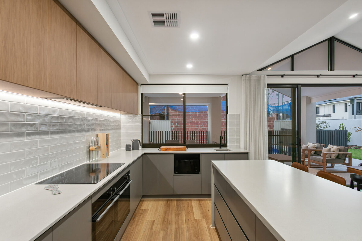 A modern kitchen with timber overhead cabinets, a white tiled splashback and an island bench, opening onto a covered outdoor area