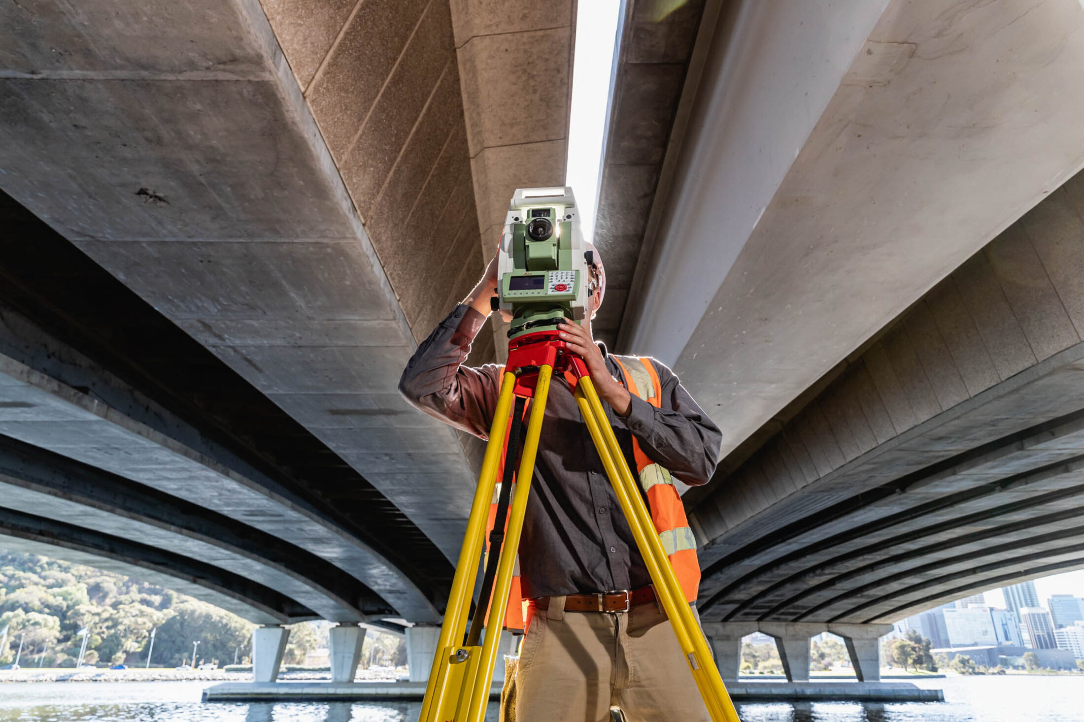 A KR Surveys surveyor using a total station on a yellow tripod under a Perth bridge